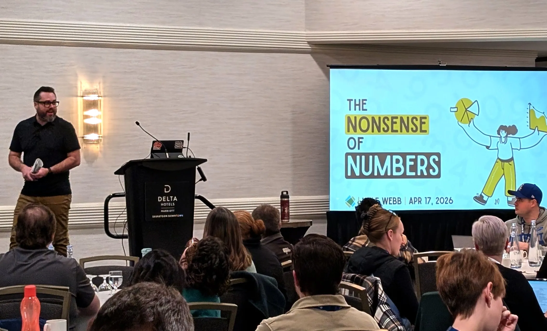 Kyle speaking to a room of teachers at round tables, beside a slide that reads The Nonsense of Numbers
